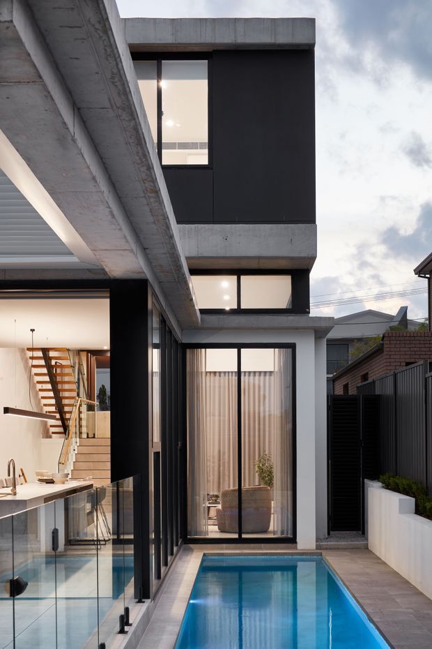 A narrow urban courtyard showing a striking contemporary multi-level house with exposed concrete cantilevered slabs, black cladding, extensive floor-to-ceiling glazing, and a compact rectangular swimming pool. The open-plan kitchen and floating timber staircase are visible through the glazing at left, while a glazed living area faces the pool. Dark metal fencing and neighbouring rooftops frame the private outdoor space.