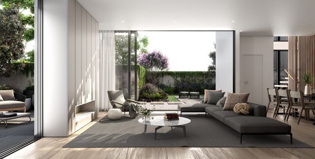 A bright, refined contemporary living room opening onto a landscaped courtyard through extensive floor-to-ceiling glazing. The room features a large charcoal sectional sofa, low round coffee tables, a neutral textured rug, vertical wall panelling, light timber-look flooring, and a dining area with timber furniture in the background. The open indoor-outdoor connection and restrained neutral palette create an airy, calm atmosphere.