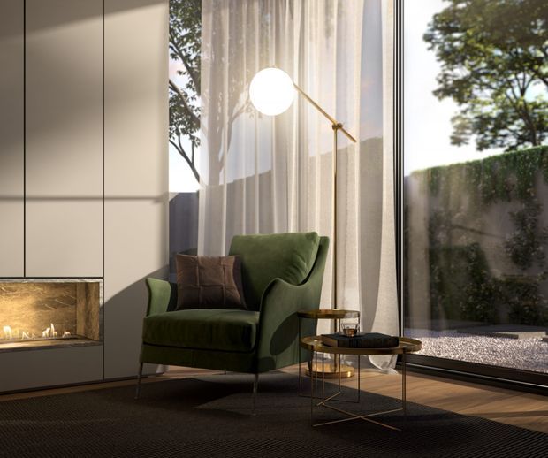 A refined contemporary living room corner with a deep green upholstered lounge chair, dark side tables, a slender brass floor lamp, sheer curtains, full-height glazing, and a built-in fireplace set into pale wall panelling. The room has warm natural light, dark flooring, and views to a private landscaped exterior.