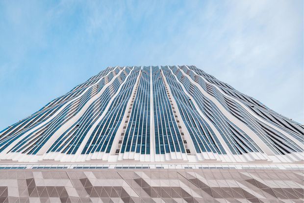 Low-angle exterior view of a large contemporary high-rise building with a broad, tapering upper facade. Dense vertical blue-tinted glazing is interwoven with white structural or cladding strips that create a rippling pattern, while the lower visible facade has geometric grey and white panels. The building rises against a pale blue sky.
