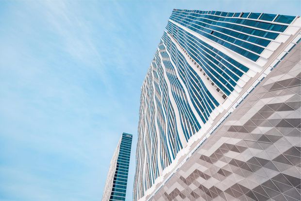 Upward-looking view of a contemporary high-rise commercial building with a sweeping, sculptural facade. The tower combines extensive blue-tinted glazing, white structural framing, and a lower or adjoining facade clad in a geometric triangular grey-and-white pattern. A second slender glass tower is visible in the background against a bright blue sky.