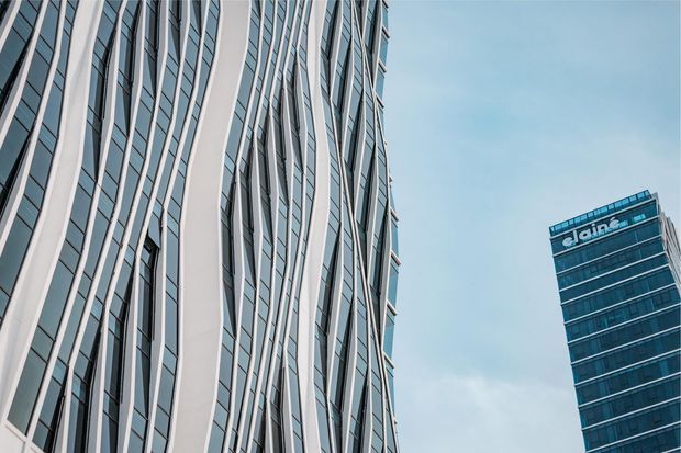 Upward-looking exterior view of two modern high-rise commercial buildings. The foreground tower has a highly articulated facade with alternating pale vertical structural bands and dark glazed window strips, while the background tower is a simpler glass-clad rectangular volume with visible signage near the roofline. The image has a cool blue-grey palette and a contemporary urban architectural character.