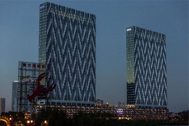 Night-time exterior view of a large contemporary commercial complex with two tall rectangular towers featuring distinctive vertical undulating light-patterned façades. A connected lower podium with illuminated glazing and signage spans the foreground, with surrounding urban elements and trees visible at the base.