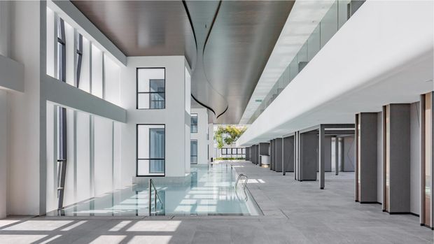 A bright, expansive contemporary architectural atrium with a shallow indoor reflecting pool or water feature, extensive floor-to-ceiling glazing, white walls, grey stone-like paving, dark vertical screens, and a sculptural curved metallic ceiling. The space appears to be a multi-storey commercial or institutional interior with an upper-level glazed balustrade and strong linear daylight.