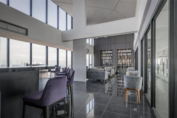 A bright, high-end contemporary open-plan interior with a bar counter and upholstered stools in the foreground, a lounge seating area beyond, and a full-height built-in shelving or display wall. Floor-to-ceiling glazing provides expansive city views, while the double-height volume, exposed structural beams, dark polished floor tiles, and neutral grey palette create a refined, spacious atmosphere.