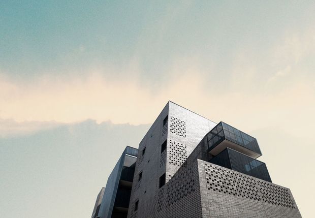 Low-angle exterior view of a multi-storey contemporary building with angular projecting volumes, dark glazed balconies, and pale masonry facades featuring extensive perforated brick or block screens. The sky dominates the background, emphasizing the building’s geometric silhouette.