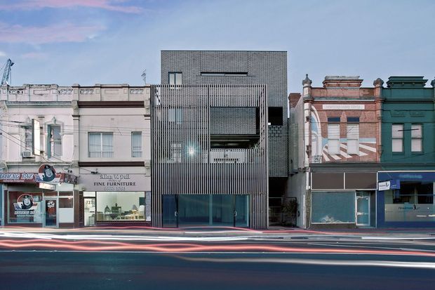 Street-level architectural photograph of a contemporary multi-storey commercial building inserted between ornate historic shopfronts. The central building has a dark grey masonry facade, vertical metal screening, recessed balconies or openings, and a glazed ground-floor frontage. Adjacent Victorian-era commercial facades feature decorative parapets and contrasting painted brickwork. Long-exposure vehicle light trails run across the foreground.