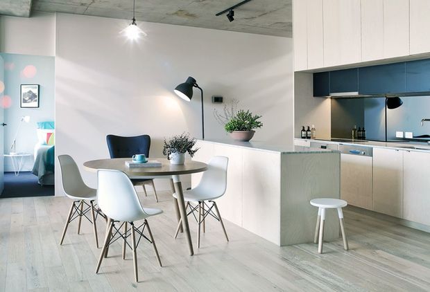 Open-plan contemporary kitchen and dining area with pale timber flooring, a round dining table surrounded by modern molded chairs, light built-in cabinetry, a dark blue kitchen splashback, and a raw concrete ceiling. A small bedroom or lounge area is visible through the opening at left.