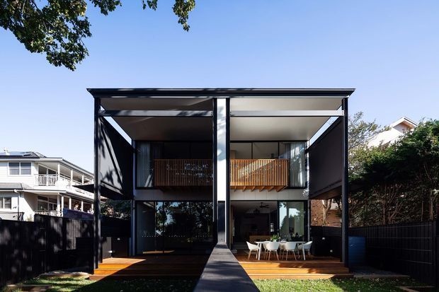 A modern two-storey residential house viewed from the front, with a strong symmetrical composition, flat roof, black structural framing, expansive glazing, recessed upper balconies with warm timber privacy screens, and timber decking on either side of a central concrete path. Fenced suburban surroundings and neighbouring homes are visible at the edges.