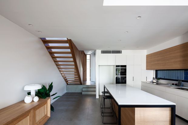 A bright, open-plan contemporary kitchen with a large white island and integrated seating, extensive white cabinetry, a timber-faced island end, and a timber overhead cabinet above the benchtop. A dark tiled floor and blue-toned splashback add contrast. An open timber stairway and hallway are visible beyond the kitchen, with a timber sideboard and indoor plants in the foreground.