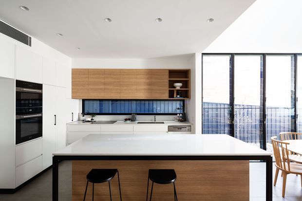 Bright contemporary kitchen with extensive white cabinetry, integrated black appliances, a large island with a white benchtop and timber-clad base, timber overhead cabinetry, blue glass-tile splashback, and full-height glazed doors opening to a balcony or terrace. A dining area is visible at the right.