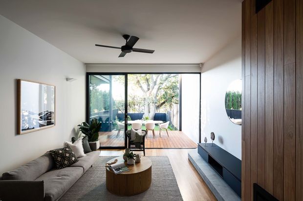 Bright contemporary living room with a grey sofa, round timber coffee table, low dark media console, and extensive floor-to-ceiling glazing opening onto a timber deck and landscaped outdoor area. A full-height dark timber feature wall and cabinetry line the right side, while a ceiling fan, indoor plants, neutral furnishings, and black-framed glazing create a refined indoor-outdoor connection.