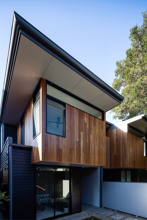 Exterior architectural photograph of a contemporary multi-level residence with projecting timber-clad upper volumes, dark blue-black wall cladding and trim, broad white soffits, expansive glazing, and a low-pitched roof. A paved entry path and boundary fencing are visible, with mature landscaping at the side.