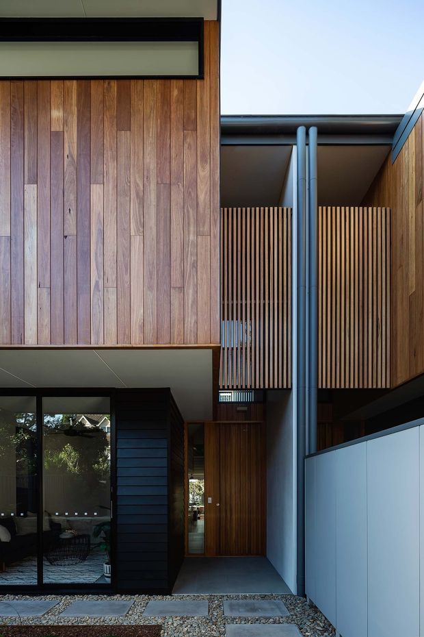 A close-up architectural view of a contemporary multi-level residential facade and entry courtyard. Vertical timber cladding and slatted timber screening frame a recessed timber front door, with dark metal framing, exposed grey downpipes, pale wall panels, and a gravel path with large stepping stones. Glazing at lower left reveals part of the interior living area. The composition emphasizes layered facade planes, privacy screens, and a sheltered entry.