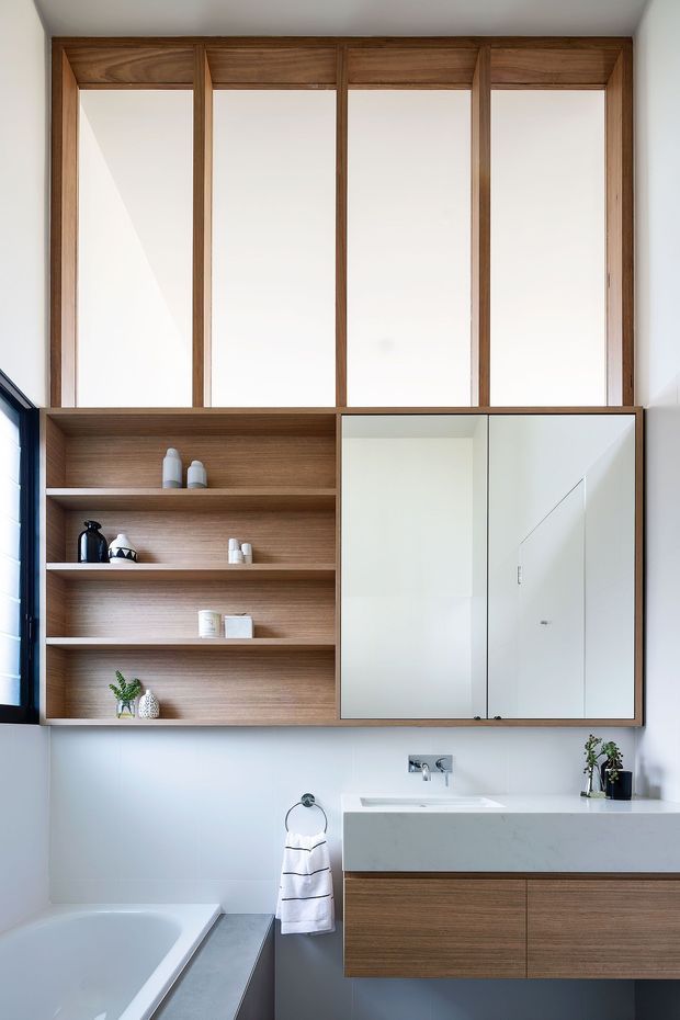 Bright contemporary bathroom with a freestanding white bathtub, floating timber vanity, white benchtop, wall-mounted basin, mirrored cabinet doors, and an open timber display niche. Large timber-framed clerestory-style glazing and white tiled walls create a clean, warm, minimalist feel.