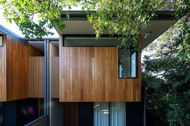 Exterior view of a contemporary multi-level house set among mature trees. The facade features warm vertical timber cladding, dark metal framing and trim, projecting box-like volumes, narrow clerestory glazing, and a flat roof with deep overhangs. Vertical timber screening adds privacy between the projecting elements.