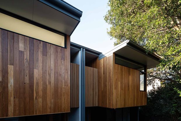 Contemporary residential exterior featuring interlocking timber-clad volumes elevated above a dark base. Vertical timber boards, slim dark metal roof edges, exposed cylindrical downpipes, recessed glazing, and a projecting flat-roofed section create a refined modern composition integrated among mature trees.