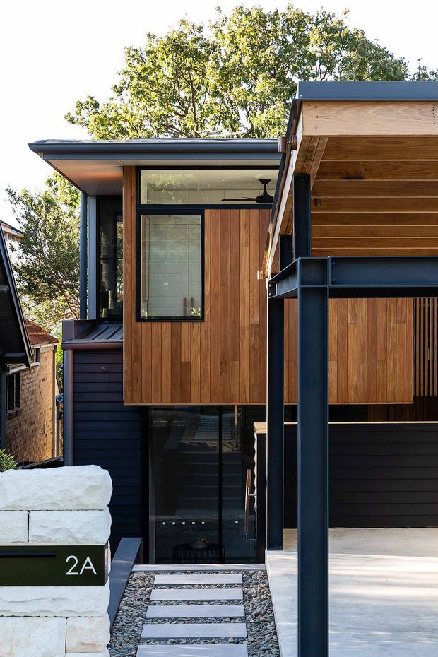 Street-facing exterior view of a compact contemporary multi-level home, featuring dark metal framing, black horizontal cladding, warm vertical timber wall panels, expansive glazing, and a projecting timber-lined carport or entry canopy. A gravel path with stepping slabs leads past a white masonry boundary wall marked 2A.