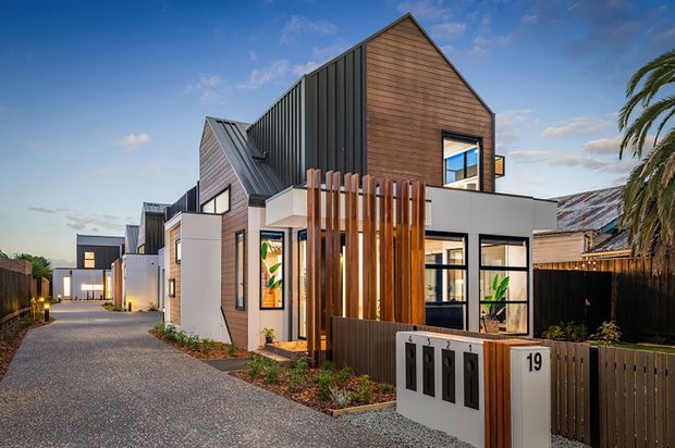 Exterior streetscape view of a contemporary multi-unit residential development. The homes feature steep gabled roof forms, dark standing-seam metal roofing, warm timber-clad upper volumes, light rendered lower levels, black-framed glazing, and vertical timber screening. A paved shared driveway and property entry signage are visible, with neighbouring houses and palm planting in the background.