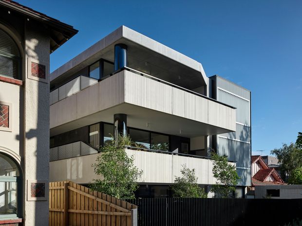 Exterior street-view photograph of a contemporary multi-storey residential building with strong horizontal balcony projections, pale vertically ribbed cladding, exposed concrete roof and balcony soffits, dark metal framing, and extensive glazing. The building sits within an established suburban streetscape beside older homes, with timber and dark metal fencing in the foreground under a clear blue sky.