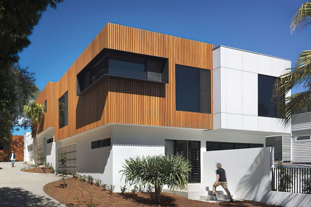 Exterior street view of a modern multi-storey residence with a strong geometric composition. The upper level is clad in vertical warm timber battens, contrasted with white rendered and white panelled volumes, dark-framed glazing, and projecting box windows. A paved pedestrian approach, landscaped garden beds, boundary fencing, and neighbouring homes are visible.