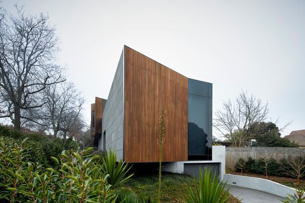 Exterior architectural photograph of a contemporary, angular single-storey building set within a landscaped suburban garden. The facade combines warm vertical timber cladding, pale grey concrete or masonry panels, and dark glazed openings. A sharply sloping roofline and projecting upper volume create a sculptural form, with a curved concrete retaining wall and paved path in the foreground.