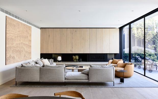 A spacious contemporary living room with a large grey sectional sofa arranged around a low timber coffee table, facing a long built-in timber and black fireplace wall. Floor-to-ceiling black-framed glazing opens toward an outdoor area, while a large textured timber-toned wall panel and neutral furnishings create a warm, refined interior.