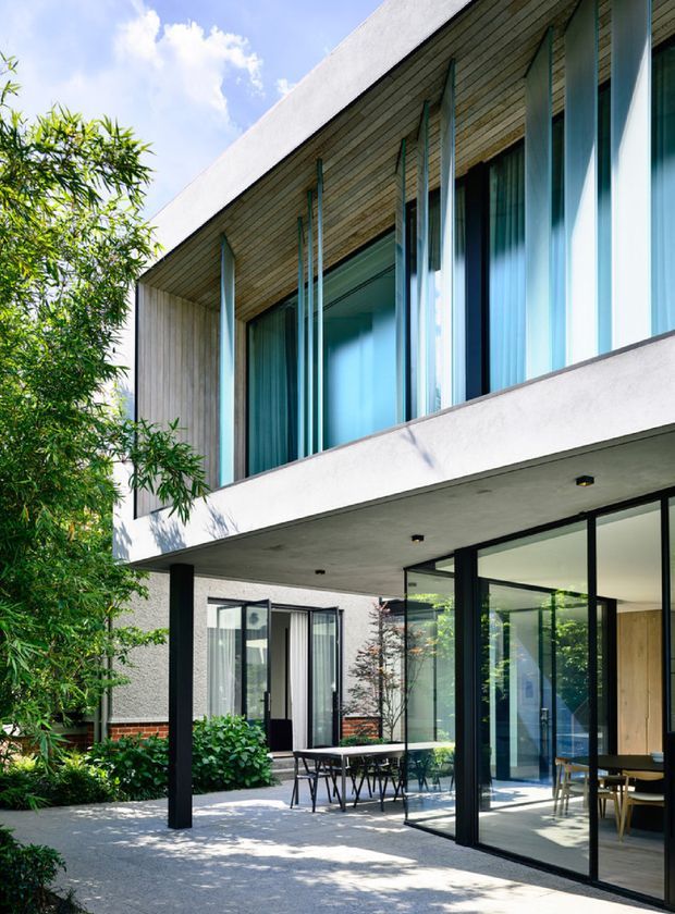 A contemporary two-storey residential exterior with a covered patio and extensive floor-to-ceiling glazing. The facade combines smooth light render, dark structural columns and frames, warm horizontal timber soffit cladding, and projecting upper-level window fins. A dining area is visible through the glass, with landscaped planting and paved outdoor space in the foreground.