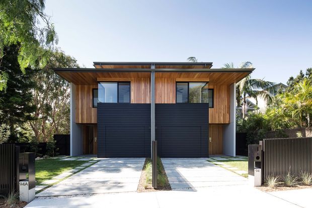 Front exterior view of a contemporary two-storey attached residential building, likely a pair of townhouses, with warm vertical timber cladding, dark horizontal garage doors, broad flat roof overhangs, concrete driveways and landscaped tropical-suburban planting. The symmetrical facade includes upper-level glazing and a central structural post, with fencing and entry pillars at the street edge.