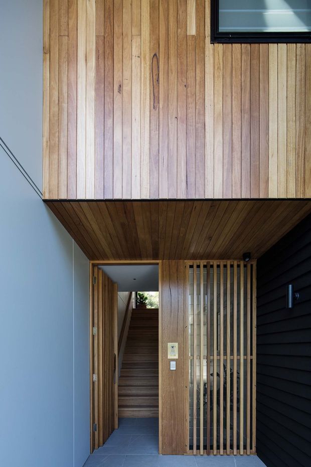 A contemporary residential entryway viewed from outside, featuring extensive warm vertical timber cladding and a timber-lined soffit. An open timber door leads to an internal stairway, while a vertical timber screen conceals the entry glazing or side passage. The entrance is framed by a pale smooth wall on the left and dark horizontal cladding on the right, with a small high-level window and grey tiled flooring.