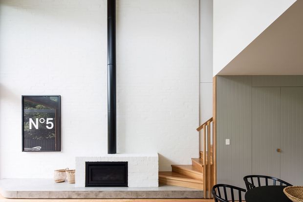 Bright contemporary interior showing a compact living and dining area with a white painted brick fireplace, black insert and flue, timber stair treads with timber balustrade, and a vertically ribbed grey built-in wall beneath a stair or mezzanine volume. Black dining chairs and a round dark table are visible in the foreground, with a framed N°5 artwork on the wall. The space has a clean, refined palette of white, timber, grey, and black.