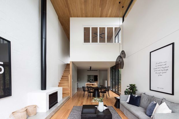 Bright double-height open-plan living and dining area with a contemporary timber-lined ceiling, white walls, a black cylindrical fireplace flue and built-in fireplace, floating timber stair, dining table, grey sofa, pendant lights, and high-level louvred windows. The space has strong vertical volume and warm natural materials.