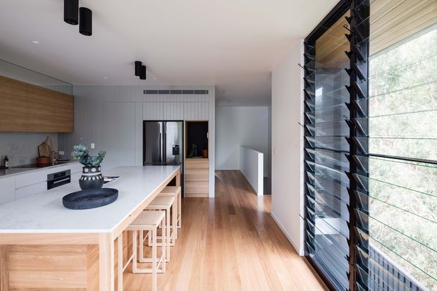 A bright, contemporary open-plan kitchen viewed from beside full-height louvred glazing. The space features a large white stone island with integrated timber seating, light timber cabinetry and flooring, a dark cooktop and oven, white walls, and black cylindrical pendant lights. A hallway and built-in storage extend beyond the kitchen, while the louvres provide filtered daylight and a strong vertical architectural feature.
