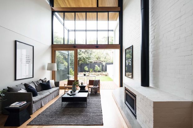 Bright contemporary living room with a large charcoal sectional sofa, black coffee table, textured rug, and white-painted brick fireplace. Full-height timber-framed glazing and glazed doors open to a timber deck and garden, while a double-height glazed upper section and exposed timber ceiling add architectural volume. Black steel fireplace flue and dark-framed artwork provide contrast against the pale walls.