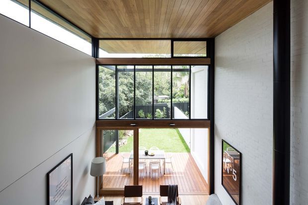 A double-height contemporary living area viewed from an elevated position, opening onto a timber deck and lawn through expansive dark-framed glazing. The room features a warm timber-lined ceiling, white painted brick walls, polished timber flooring, and a tall black cylindrical fireplace or flue. Dining and seating furniture are visible below, with framed artwork on the walls and a leafy garden beyond.