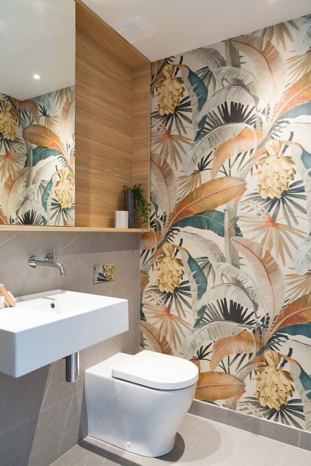 Compact contemporary bathroom featuring a wall-mounted white basin, chrome wall-mounted tap, concealed-cistern toilet, large mirror, and timber-look built-in cabinet. Grey tiled surfaces contrast with a bold tropical botanical feature wallpaper in muted green, cream, beige, terracotta and charcoal tones.