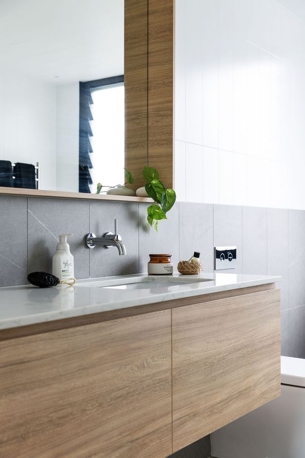 A bright, contemporary bathroom vanity viewed close-up. The room features a floating light timber vanity with integrated drawers, a pale stone or engineered-stone benchtop and basin, large light grey wall tiles, a chrome wall-mounted tap, a large mirror, and glossy white upper wall tiles. Minimal toiletries and a small trailing plant add restrained decorative detail.