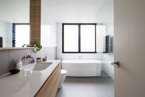 Bright contemporary bathroom viewed from the doorway, featuring a floating timber-look vanity with a white stone benchtop and integrated basin, wall-mounted tapware, large-format pale grey floor and wall tiles, a freestanding white bathtub, frameless glass shower screen, black-framed louvred window, and a full-height mirror. Minimal decor and indoor plants add subtle warmth.