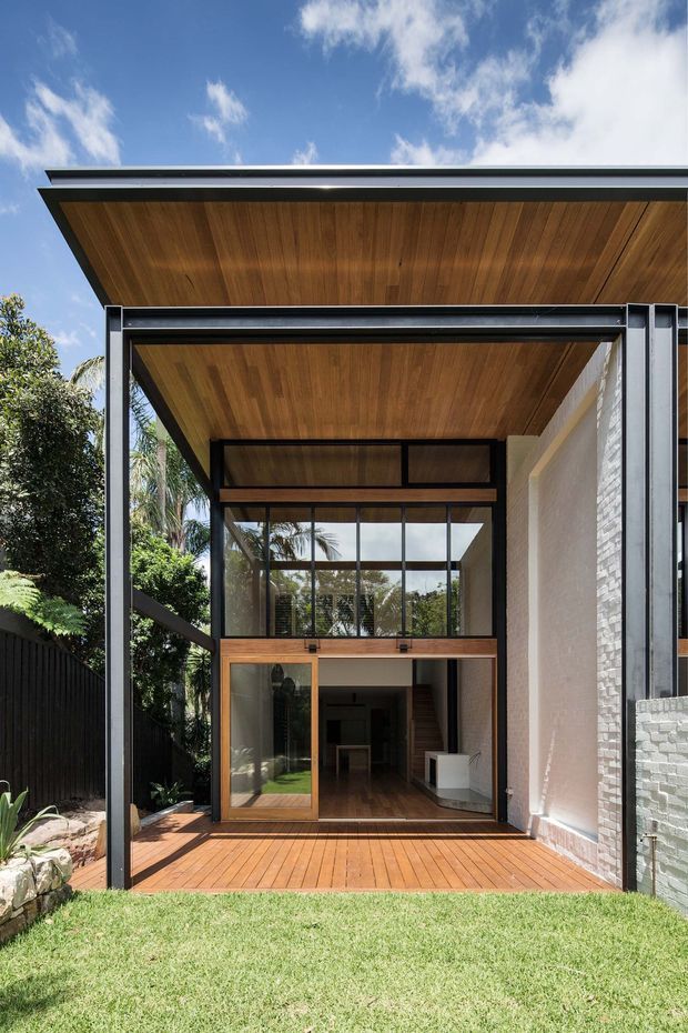 Exterior view of a contemporary two-storey glazed extension or courtyard-facing facade. The structure features a broad flat roof with warm timber soffit lining, slim dark metal framing, extensive floor-to-ceiling glazing, a timber-framed glazed door, and a timber deck. A white painted masonry wall forms the side elevation, with landscaped lawn and tropical vegetation surrounding the building.