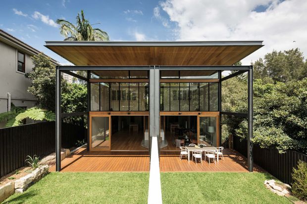 A contemporary two-storey rear extension or pavilion viewed from the garden, featuring extensive black-framed glazing, warm timber soffits and decking, and a broad low-pitched roof. The glazed lower level opens onto a timber deck with outdoor dining furniture, while the upper level provides additional enclosed space. The structure is set within a landscaped suburban garden.