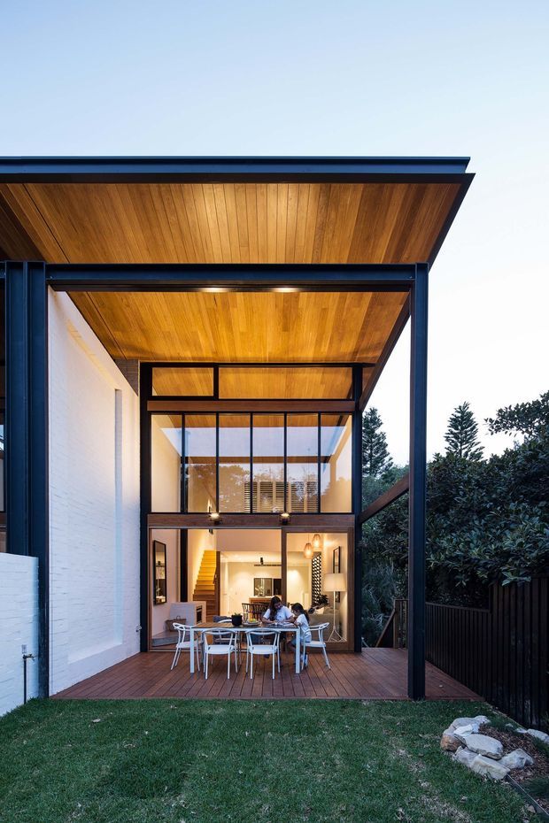 A rear exterior view of a contemporary two-storey home opening onto a timber deck and lawn. The design features a large covered outdoor dining area, black steel framing, extensive glazing, white masonry walls, and a warm timber-lined soffit and ceiling. Interior living and dining areas are visible through the glazing, with a stairway and upper-level void apparent inside. Dense planting and dark fencing frame the residential courtyard setting.