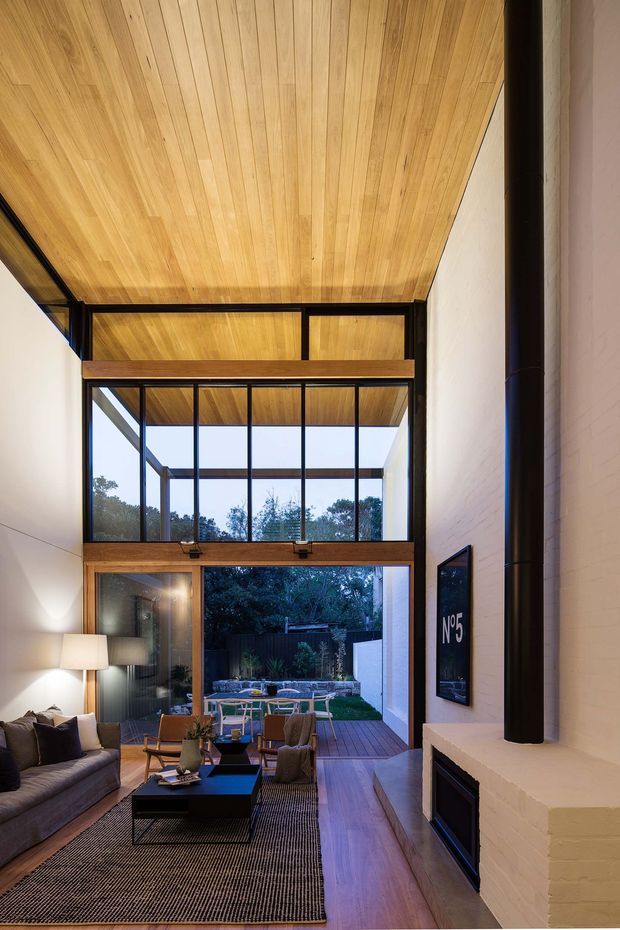 A tall, narrow contemporary living room with a dramatic double-height timber-lined ceiling and large black-framed glazed doors opening to a timber deck and landscaped courtyard. The room includes a grey sofa, armchairs, low black coffee table, patterned rug, fireplace with a dark vertical flue, and warm timber detailing. The glazing and ceiling create a strong indoor-outdoor connection with abundant natural light.