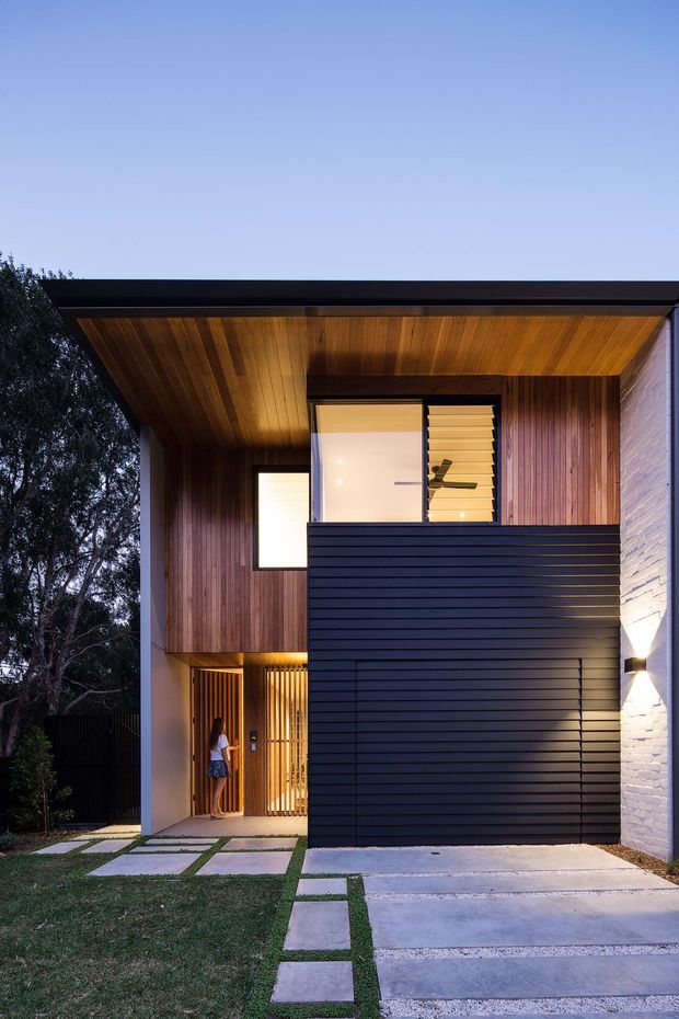 Street-facing exterior view of a contemporary multi-level home at dusk. The facade combines dark horizontal cladding, warm vertical timber cladding, a timber-lined recessed entry canopy, a white-painted brick side wall, and large windows with external louvres. A partially enclosed timber-screened entry sits beside a concrete driveway and stepping-stone path through the lawn.