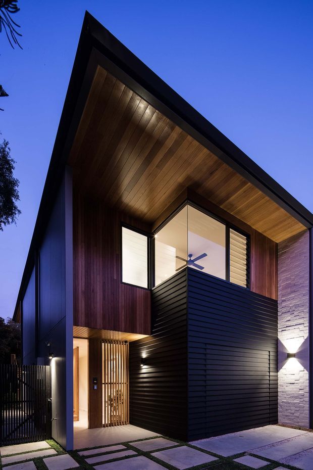 Twilight exterior view of a tall, sharply angular contemporary residence. The facade combines dark vertical timber cladding, black horizontal metal-like cladding, extensive glazing, a warm timber-lined projecting soffit, and a pale textured masonry wall. A recessed illuminated entry, slatted privacy screen, balcony enclosure, and large-format paved forecourt are visible.