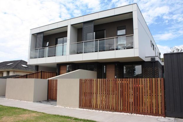 Street-facing view of a contemporary multi-storey townhouse or apartment-style residence. The building has a crisp white rendered facade, dark brickwork at ground level, projecting upper-floor balconies with clear glass balustrades, timber entry doors, vertical timber fencing, and dark louvred sun-shading panels. A neighbouring residential roof and landscaped verge are visible.