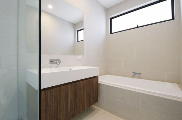 Bright contemporary bathroom featuring a wall-mounted white vanity with dark timber cabinetry, a large frameless mirror, glass shower screen, tiled walls and floor, and a built-in bathtub beneath a narrow horizontal window.