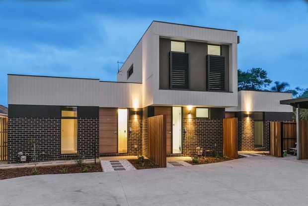 Exterior view of a contemporary multi-level residential development at dusk, featuring a rectilinear rendered upper volume, dark brick ground-level walls, vertical timber screening and gates, narrow windows with dark louvred shutters, and integrated warm exterior lighting. A broad concrete driveway or forecourt occupies the foreground.