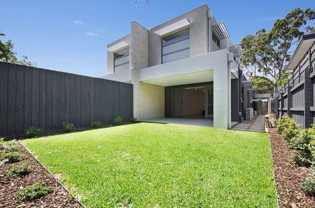 Exterior view of a contemporary multi-storey residence with a crisp white and grey facade, projecting box forms, broad glazing, and horizontal sun-shading elements. The home fronts a manicured lawn bordered by dark timber fencing and landscaped planting, with a covered entry/garage-like opening beneath the upper level.