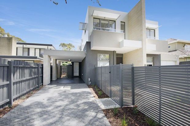 Exterior view of a contemporary multi-storey residential townhouse with white rendered walls, projecting balconies, flat roof forms and screened private front areas. A paved driveway leads toward a covered entry or carport, with dark horizontal slatted fencing and neighbouring homes visible.