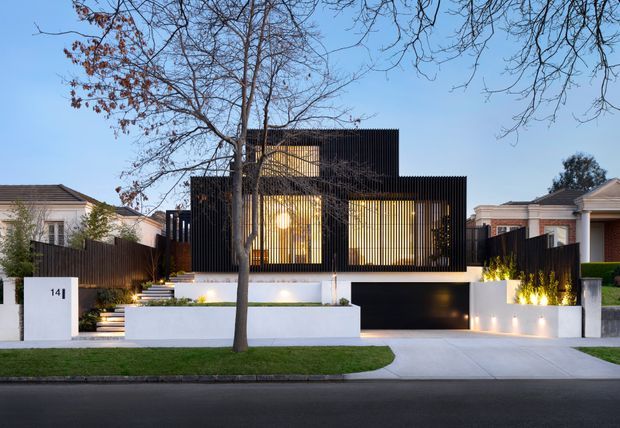 Street-facing view of a contemporary multi-level residence with a strong geometric composition, black vertical screening fins, warm illuminated glazing, white rendered walls, a recessed dark garage, landscaped front garden, and broad concrete driveway. The house is set among neighbouring suburban homes beneath mature street trees.
